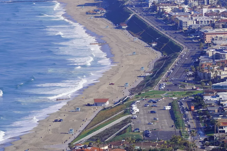 A parking lots and homes sit on a bluff above a large beach on Pacific coast in Southern California.